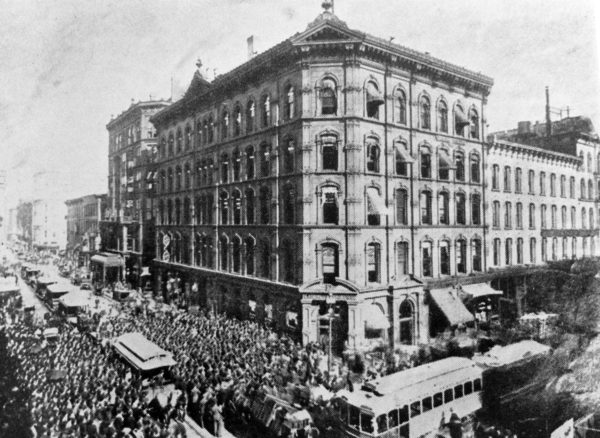 A historical black and white image of six story Italianate corner building with heavy traffic in front.