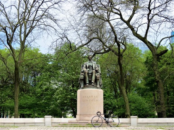 A tour bike standing in front of the pedestal on which sits a seated Abraham Lincoln statute with green trees and a skyscraper in the background.