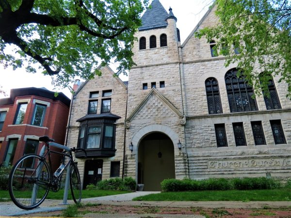 A tour bike standing in front of a white limestone Romanesque church and an attached later rectory with a dark brown wood bay window.