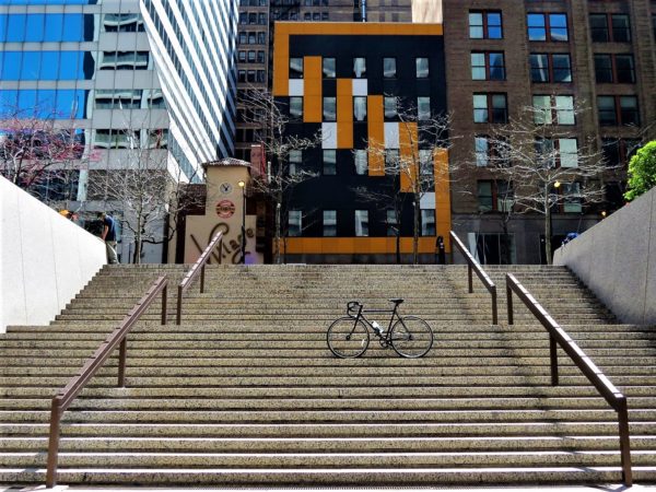 A tour bike standing in the middle of two flights of stone steps with a modern orange and glass city building in the background