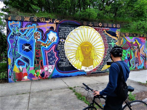 A CBA bike tour rider taking a picture of Afro-futuristic mural demoniated by a Sun like human head.