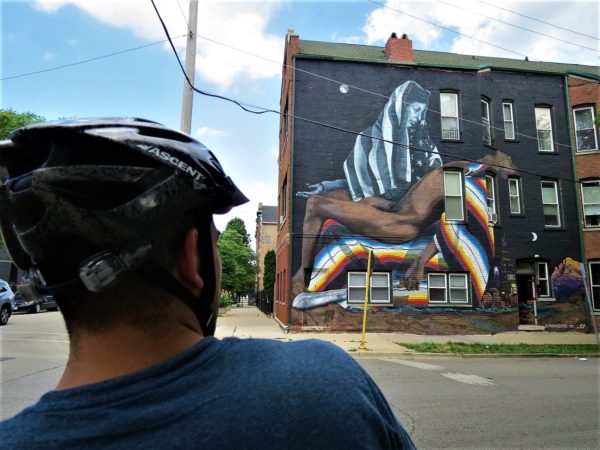 A CBA bike tour rider looking at an La Pieta style mural of a Virgin Mary figure with an American flag head covering and a brown skinned Christ figure draped in an indigenous blanket laying in her lap filling an entire building side.