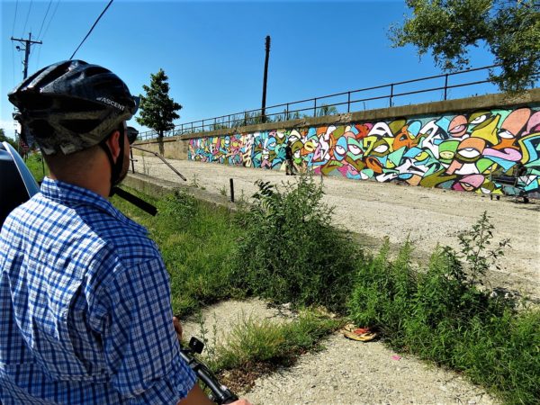 A CBA bike tour rider watching a street artist working on a mural of multicolored interlocking shapes that can be read by the experienced