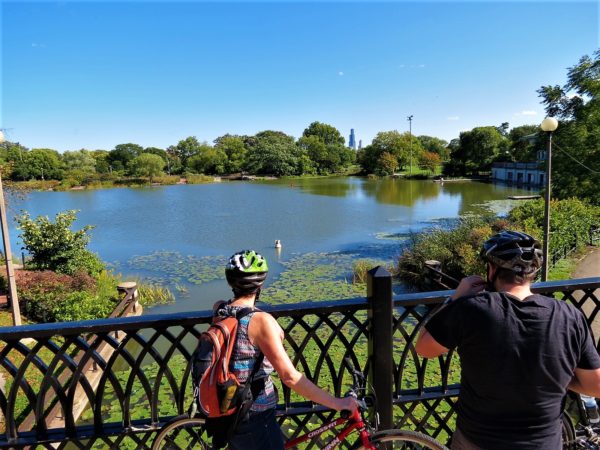 Two CBA bike tour riders on a bridge overlooking a park lagoon surtounded by greenery with the Willis Tower in the distance