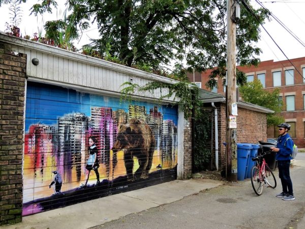 A CBA bike tour rider taking a picture of a garage mural of a pegion, girl, and bear walking single file in front of a dreamlike colorful city.
