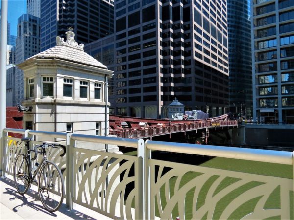 A tour bike leaning on a chrome railing to the side of a maroon painted movable bridge and its white limestone bridge house with skyscrapers in the background,