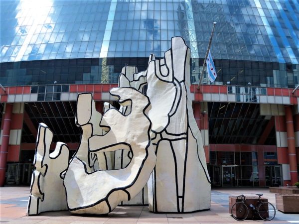 A tour bike standing to the side of a white black outlined abstract sculpture with a modern class and red steel building in the background