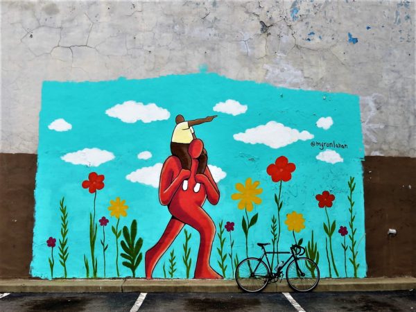 A tour bike leaning on a mural of a faceless brown skin figure with another smaller figure on its shoulders pointing into the distance whlie they walk among flowers.