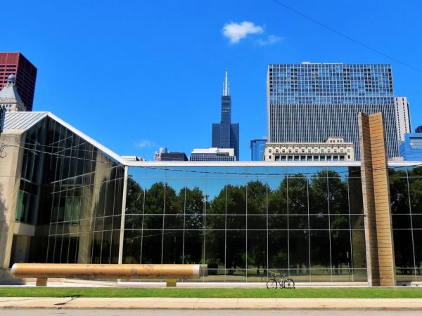 A tour bike standing between earth colored cylinder and and tower sculpture with a three story glass wall behind and skyscrapers in the background.