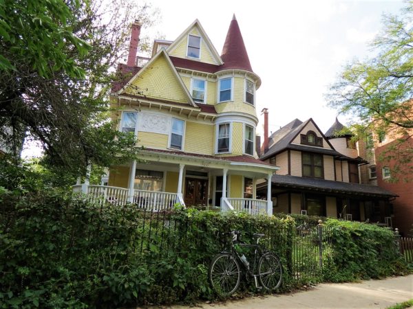 A tour bike leaning on the green hedge in front of a three story yellow painted wood Victorian home with a pointed corner turret.