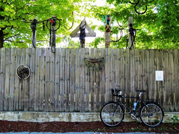 A tour bike leaning on a wooden fence with found bike parts art installed.