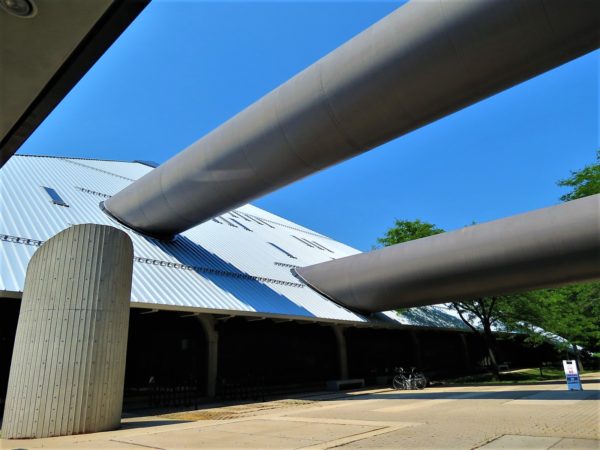 A tour bike leaning below two second and third floor dark grey tubes emerging from the above right and entering a steel pyramid building at the left.