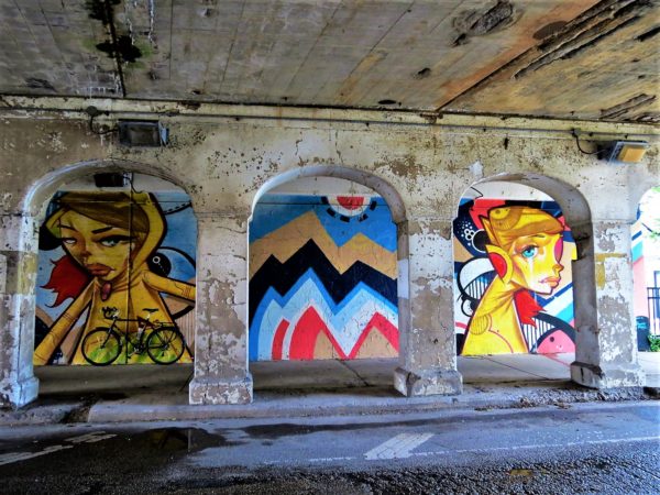 A tour bike leaning on a mural with two painted figures and a colored pattern framed by the flaking white painted arches of an underpass.