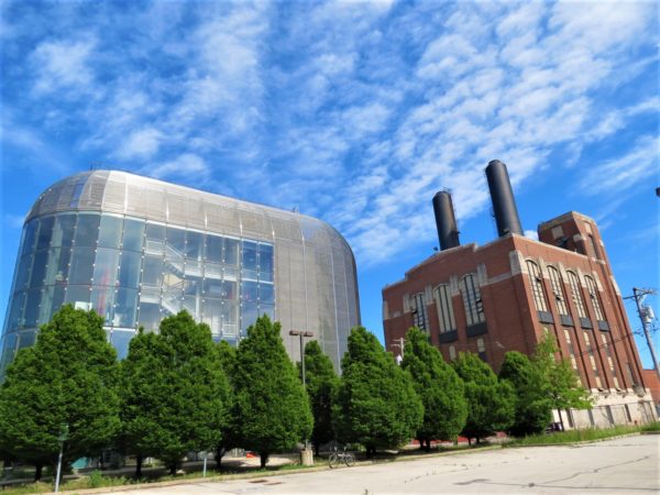 A tour bike standing in front of a bank of trees in front of two utility buildings, on the left a modern steel and glass box five story and the right a red brick and terra cotta detailed box five story with two smokestacks.