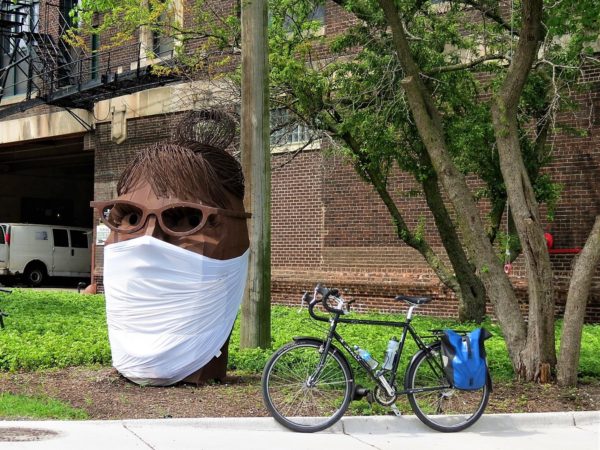 A tour bike standing next to a dark brown metal screaming Sarah Palin head with a face mask covering her mouth.