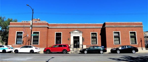 A tour bike at the curb between parked cars in fornt of a art deco neo classical red brick and stone one story postal building