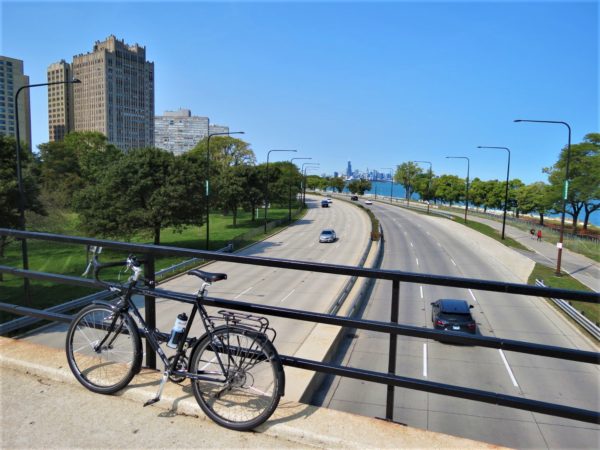 A tour bike leaning on the bridge railing over a six lane road with an apartment tower and Chicago skyline in the distance.