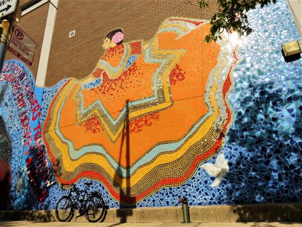 a tour bike leaning on a wall mosaic of a woman in a billowing orange traditional a Mexican dress.