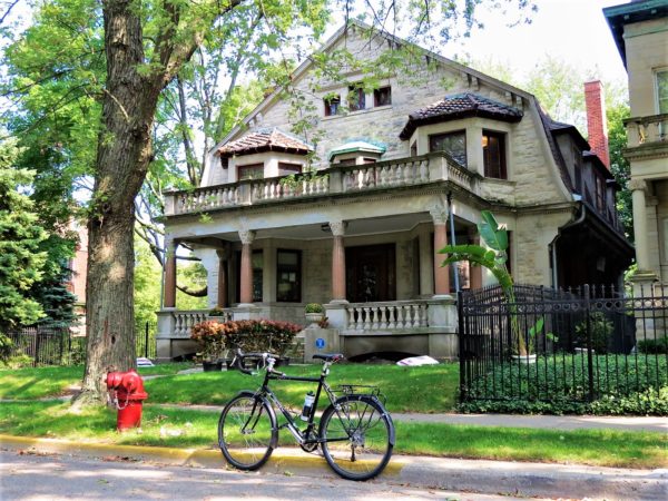 A tour bike standing in front of a three story limestone faced three wide three story home with a stone front porch and three second story only bay windows.