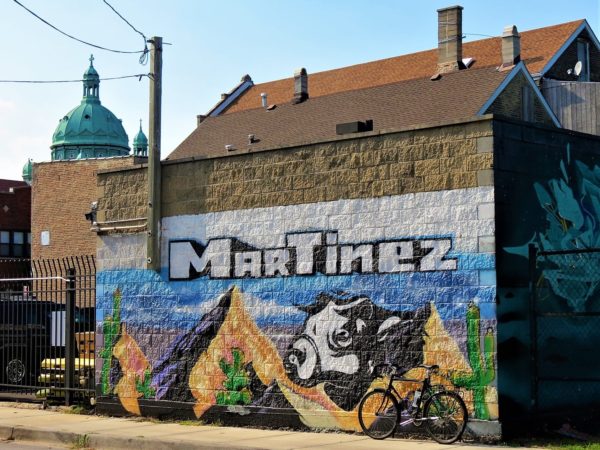 A tour bike leaning on a commercial mural for Martinez Grocery depicting a cows head looming over three mountain tops with three green copper domes of a church in the background.