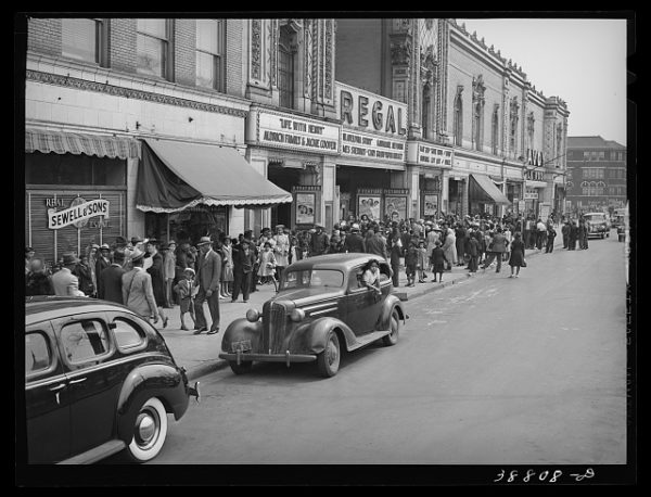 A historical black and white of 1940s cars and people in front of a lighted movie theater.