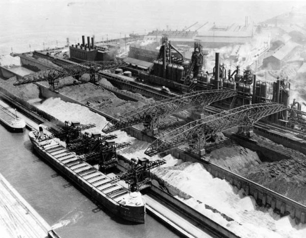 A historical black and white of a large steel plant with three steel rail bridges spanning raw materials and ending at a waiting ship.