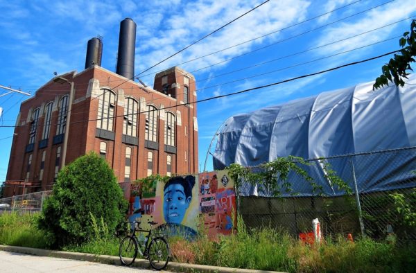 A tour bike standing a the curb next to a tarp covered garden building, a painted wooden panel and an early 20th century red brick industrial 8 story block mass building with two black smokestacks.