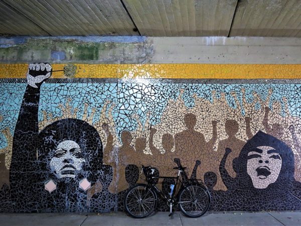 A tour bike leaning on a mosaic of two woman, one Black with an afro and a raised fist, the other Latina in a beret, with the outlines of a rising crowd between them,