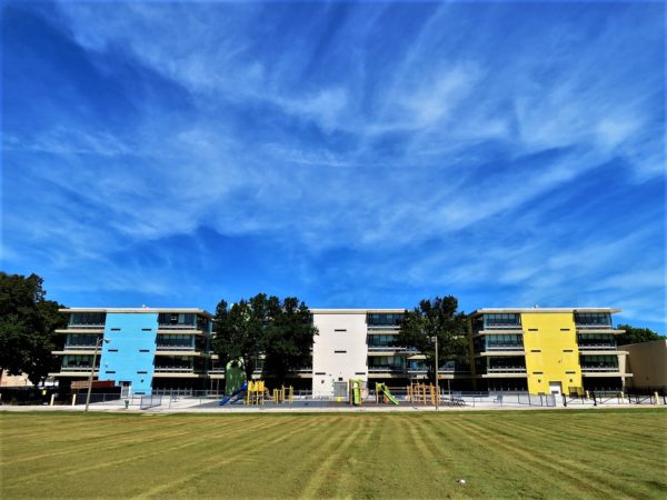 A barely visible tour bike leaning on a distant chainlink fence in front of a three section, bl Mid Century Modern school with freshly mowed grass and a wispy clouded blue sky.