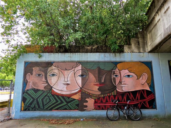 A tour bike leaning on a mural of four ethnically diverse faces merging in a Cubist way to make more faces.