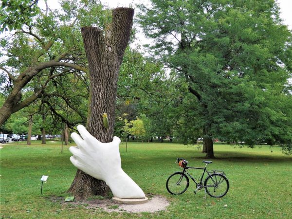 A tour bike standing next to a sculpture of a white hand emerging from the ground to grasp the limbless trunk of a tree just below a large bug