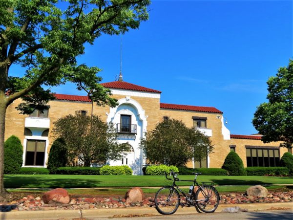 A tour bike standing at the curb in front of a Spanish Revival tan brick and red roof building with manicured bushes in front.