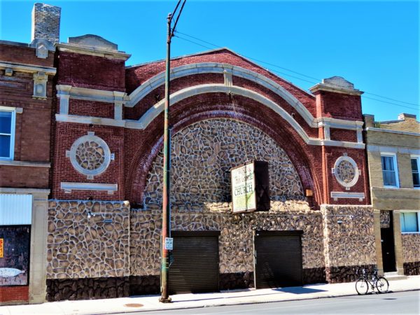 A tour bike in front of a red brick and faux stone auditorium building with whotwe limestone details and two brown door entries under a wide stone filled arch.