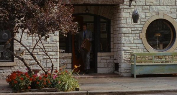 Will Ferrell walking out of a door next to an ocular window in a stone covered wall.