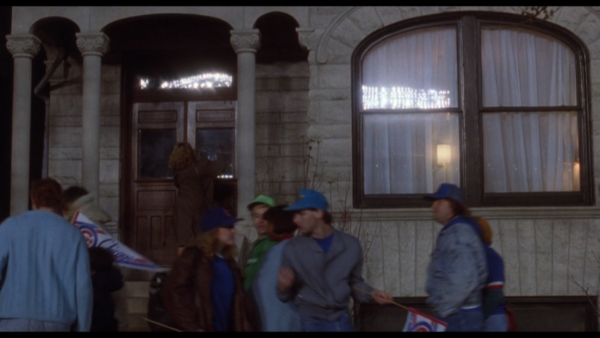 A movie still of people wearing Cubs baseball hats walking past a greystone front window.