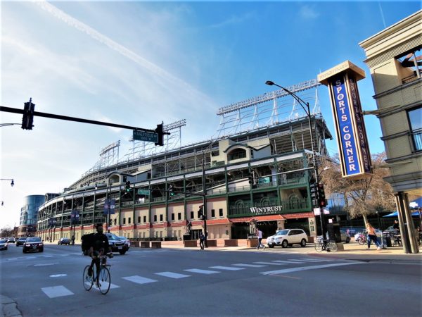 A bicyclist riding towards the viewer with a tour bike standing at the corner in front of Wrigley Field and a sign that reads Sports Corner.