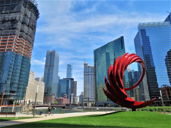 A red swirl sculpture and bike tour rider with a back drop of a river running through skyscrapers.