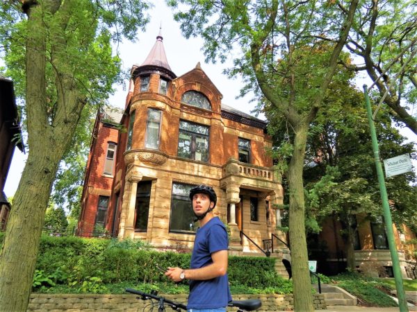 A bike tour rider looking away from the camera with a red brick and sandstone Queen Anne three story home with a cone topped corner turret.