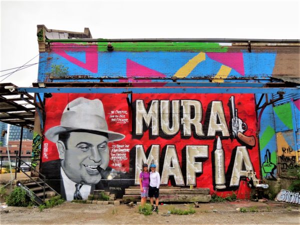 Two bike tour riders standing in fornt of a red background mural with a cartoon black and white Al Capone with bubble letters hat say Mural Mafia