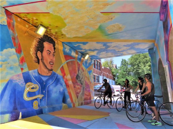 Four bike tour riders in an underpass entirely covered in a mural of a baseball player amongst sun and clouds