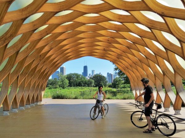 Two bike tour riders underneath a wooden honey comb-like tunnel with the Chicago skyline in the distance