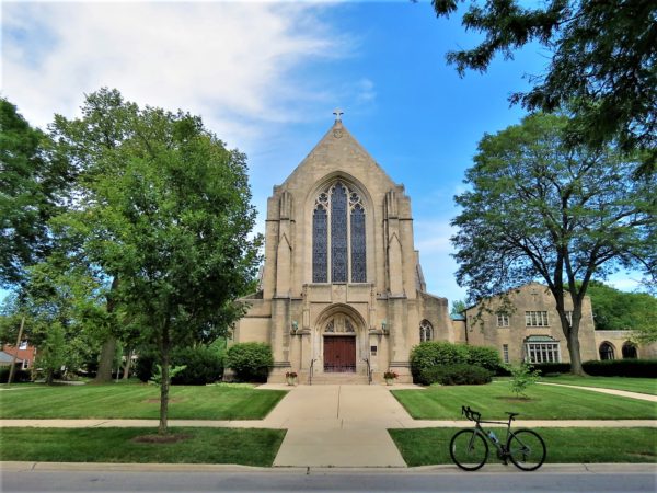 A tour bike at front of a grey limestone Gothic Revival towerless church with a large center arched window.