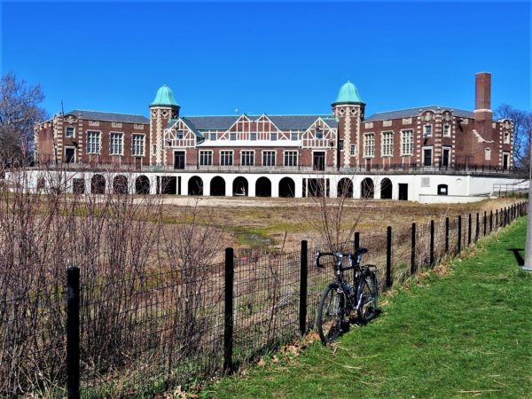 A tour bike leaning on a black wire fence with a Tudor Revival park building with two coppper domed three story towers in the background.
