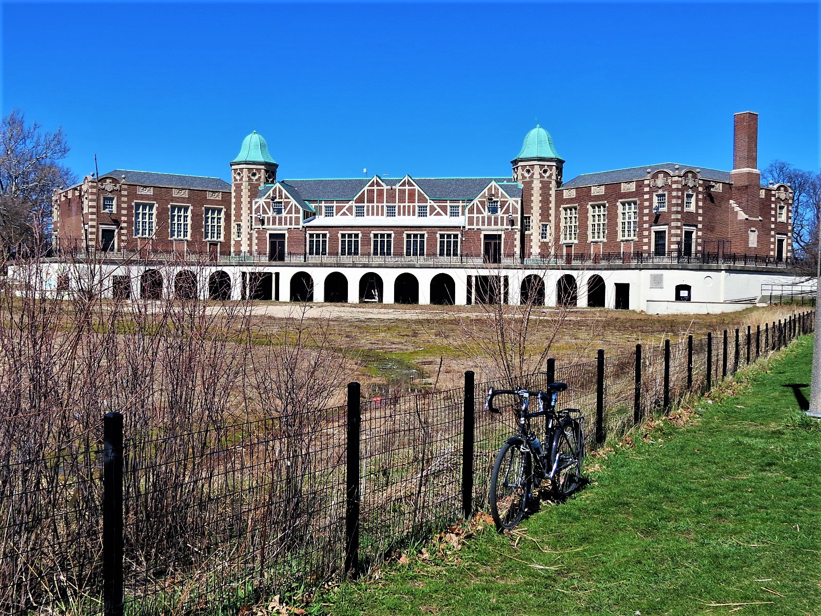A tour bike leaning on a black wire fence with a Tudor Revival park building with two coppper domed three story towers in the background.