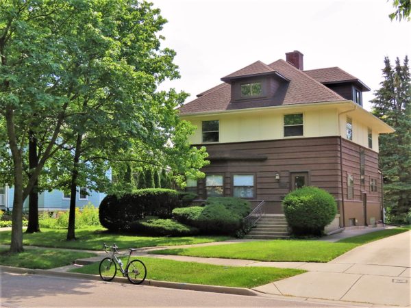 A tour bike at front of a two and half story box home with wood paneling up the the first and half of the second floor and hipped roof.