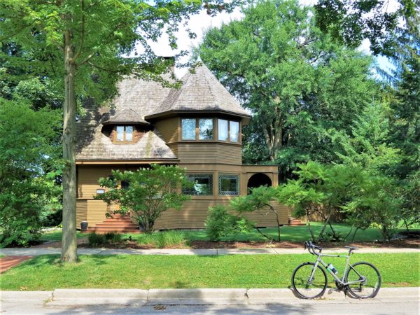 A tour bike at front of a brown painted wood Queen Anne style home surrounded by green trees.