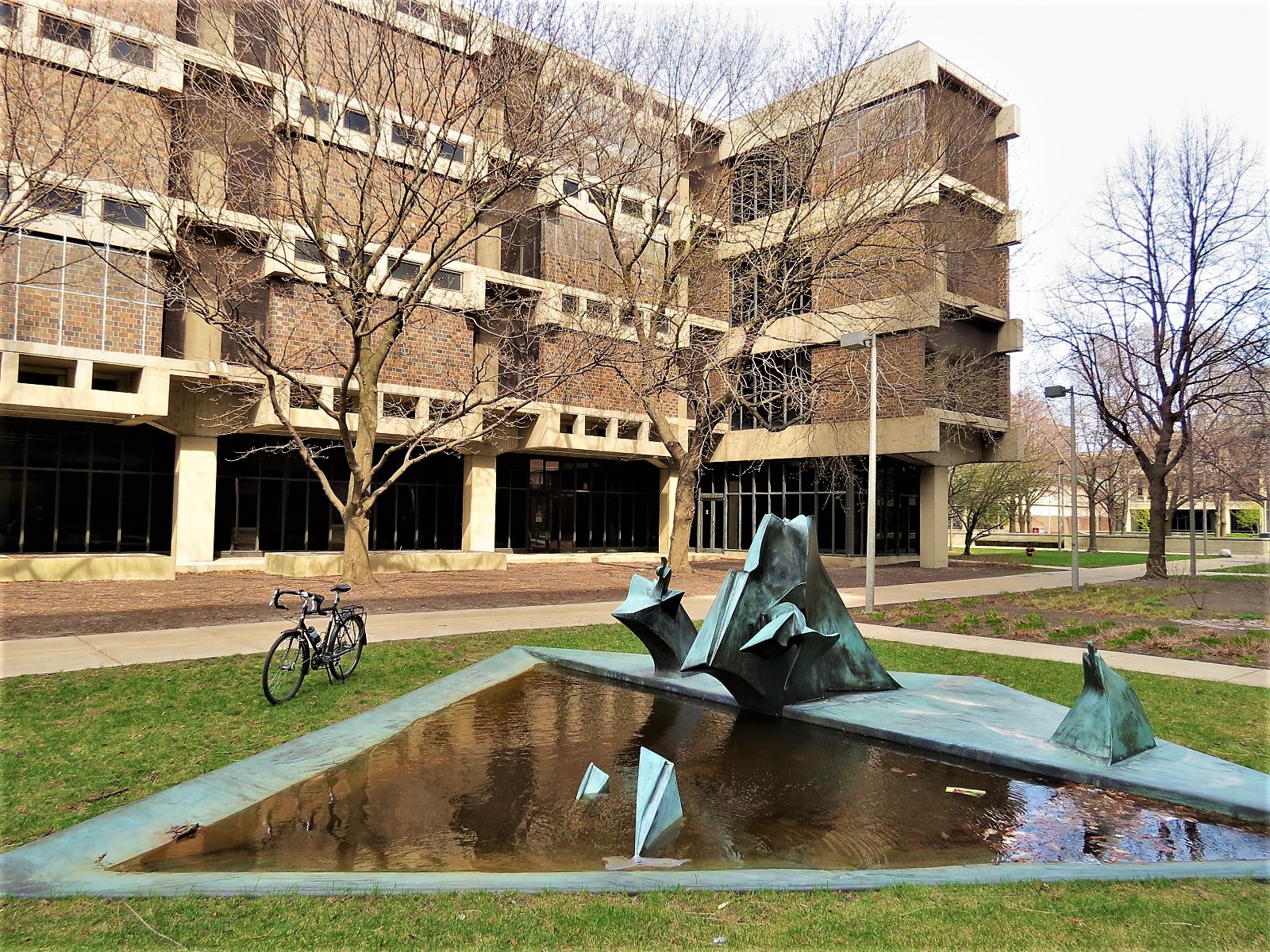 A tour bike standing behind a green patina abstract sculpture pond with four story Brutalist building behind.