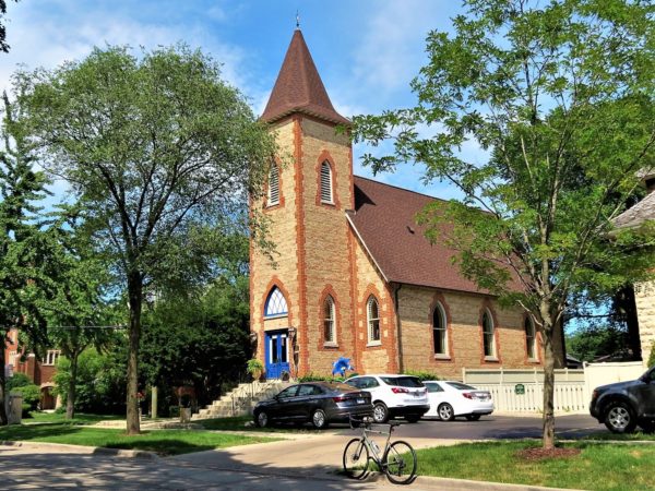 A tour bike standing to the side of a tan brick with red brick detail church with a three story square entrance tower.