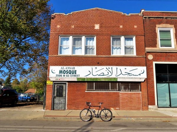 a tour bike at front of a red brick two story with a green on white banner over the first floor windows and door that read Al-Ansar Mosque in English and Arabic.