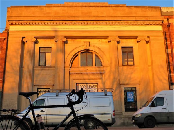 A tour bike in silhouette with a limestone Classical Revival two story former bank behind, bathed in golden hour light.